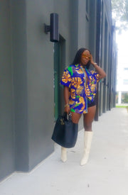 Beautiful model posing in bold purple Hawaiian Menswear shirt top accented with orange leaves in floral print , black tights,  white boots,  caramel sunglasses and black purse